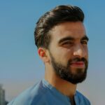 A close-up outdoor portrait of a young man with facial hair, exuding confidence under a clear sky.